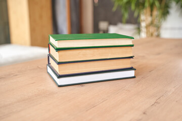Stack of hardcover books on wooden table in cozy indoor setting with greenery and natural lighting.