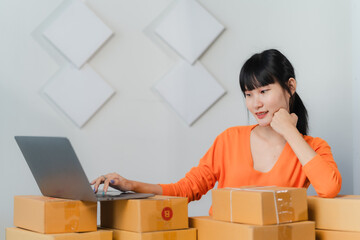 Online Business Owner: Asian woman managing orders on laptop among cardboard boxes 