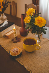 Cozy dining table with a warm autumnal atmosphere featuring a yellow cup, an open book, a muffin, and decorative vases with yellow flowers. The scene evokes a sense of relaxation and comfort