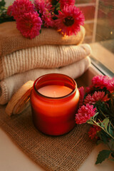 Autumn cozy still life with aromatic candle in glass jar, pink flowers and soft knitted sweaters