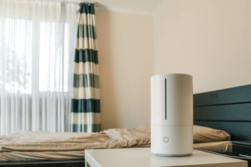 White humidifier on a bedside table near a bed with beige bedding. Concept of home comfort, air quality, and healthy sleep environment
