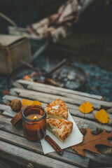 Close-up of a lit candle in an amber glass jar, two slices of apple pie, and a cinnamon stick on a rustic wooden table. Autumn leaves and walnuts enhance the cozy atmosphere near a warm campfire