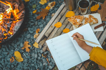 Person writing in a notebook next to a bonfire during autumn. Outdoor cozy setting with fallen leaves, snacks, hot drink, and the warmth of the fire, capturing the essence of a peaceful autumn day