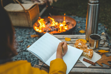 Person writing in a notebook next to a bonfire during autumn. Outdoor cozy setting with fallen leaves, snacks, hot drink, and the warmth of the fire, capturing the essence of a peaceful autumn day