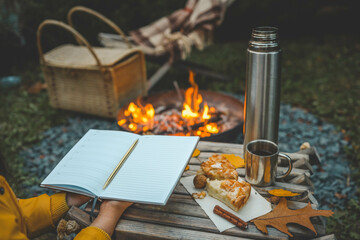 Person writing in a notebook next to a bonfire during autumn. Outdoor cozy setting with fallen leaves, snacks, hot drink, and the warmth of the fire, capturing the essence of a peaceful autumn day