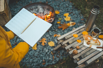 Person writing in a notebook next to a bonfire during autumn. Outdoor cozy setting with fallen leaves, snacks, hot drink, and the warmth of the fire, capturing the essence of a peaceful autumn day
