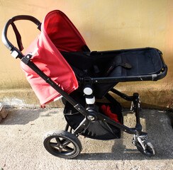 A fashionable red-black empty baby carriage in a garden in front of the yellow house.