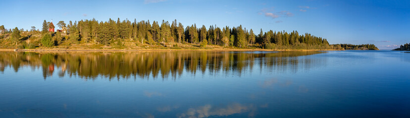 Wide-angle panorama of a tranquil sea with reflections of a dense forest and distant red lighthouse under a clear blue sky, creating a peaceful atmosphere.