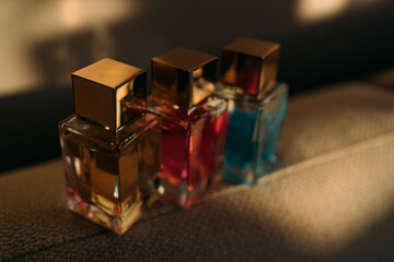 three glass perfume bottles with gold caps in yellow, pink, and blue shades on textured surface, with soft shadows in warm, ambient lighting