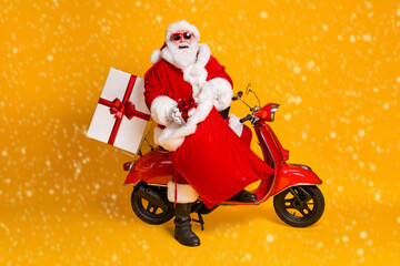 Full body profile side photo of grey white hair bearded santa claus sit scooter put gift box in sack wear red headwear sunglass boots isolated bright shine color background