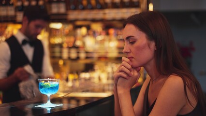 Sitting beautiful woman guest feeling bored waiting friends or couple with drinking mixed vodka in blue hawaii cocktail at luxurious counter bar for Friday meeting on bartender background. Vinosity.