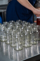 set of empty glass jars lined up on a stainless-steel table in an artisan jam factory. A worker is preparing the jars for filling with handcrafted rosehip jam. 