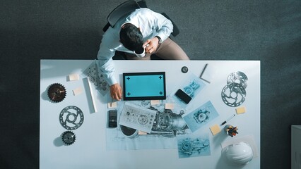 Top down aerial view of engineer hold tablet while learning about engine. Aerial view of skilled project manager looking at tablet while sitting at table with engine and drinking coffee. Alimentation.