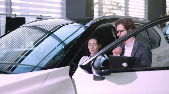 A businesswoman sits behind the wheel of a new car, ready for a test drive, while a salesman offers insights. This moment captures the excitement of choosing the perfect vehicle for her needs.