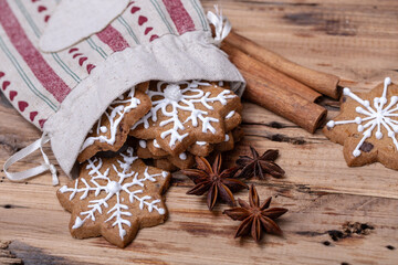 Christmas-themed gingerbread cookies with white icing snowflake designs spilling from a festive cloth bag, surrounded by star anise and cinnamon sticks on a rustic wooden surface.