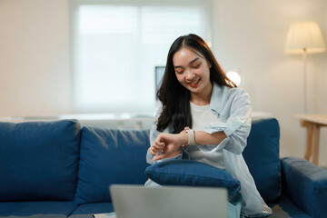 Young woman is sitting on a blue sofa and checking the time on her smartwatch while working remotely on her laptop