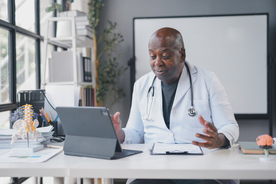Senior doctor is sitting at his desk and having a video call on his tablet, gesturing with his hand as he talks