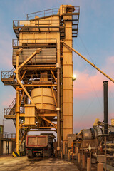 Dump truck under an asphalt manufacturing plant waiting for the mixing process of bitumen and gravel to finish in the silo.