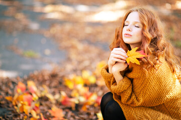 A serene woman with red hair holds an autumn leaf close to her face, eyes closed, wearing a cozy...