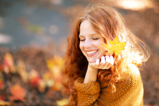 A smiling woman with red hair holds an autumn leaf near her face, eyes closed, enjoying the warmth of the sun, surrounded by fallen leaves in a serene outdoor autumn setting.