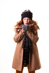 Woman with red hair, wearing a black beanie, gray gloves, and a brown coat, blows into her hands to warm them, with snow on her clothing and hat, standing against a white background.