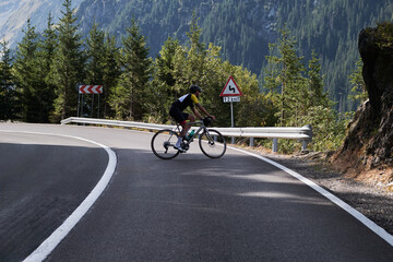 Cyclist riding a bike up a winding mountain road surrounded by lush forest. A sign warns of a bend...