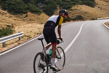 Fototapeta premium Rear view of a cyclist riding on a winding road through the mountains. Man cyclist riding a gravel bike with a view of the mountains. Travel biking concept. Transfăgărășan road.