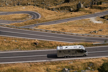 Aerial view of a motorhome driving on a winding mountain road. The road snakes through the landscape, with grassy hills and a clear blue sky overhead.