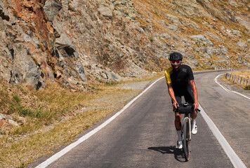 A cyclist in a yellow jersey and black shorts rides a bike on a mountain road. He wears a helmet and sunglasses and is smiling.