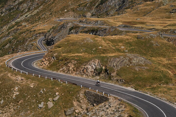 A lone cyclist rides on a winding mountain road, with scenic views of grassy hills and rocky...