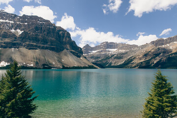 view of a rocky cliff with snow patches next to a calm lake with fluffy clouds in the sky in summer