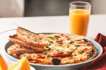 Frittata or Italian omelette with ham, tomatoes, cheese on a blue plate with slices of toasted bread. A glass of orange juice and a burgundy napkin with cutlery. Selected focus.