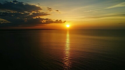 Naklejka premium A stunning sunset over the ocean with a bright sun and golden light reflected on the water. The sky is a mix of orange, yellow, and blue with dramatic clouds.