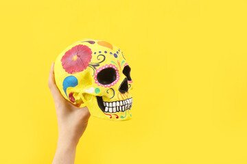 Female hand holding painted human skull for Mexico's Day of the Dead (El Dia de Muertos) on yellow background