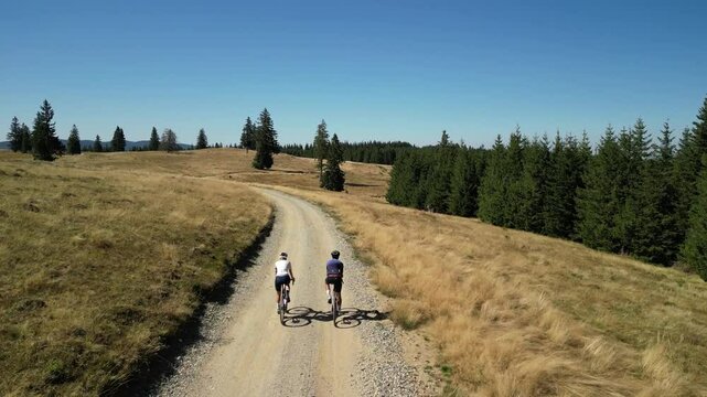 Two cyclists ride on a gravel road in a scenic mountainous landscape. The sun shines brightly, casting long shadows on the path. .Cycling on gravel roads or off-road. Active cycling adventures .