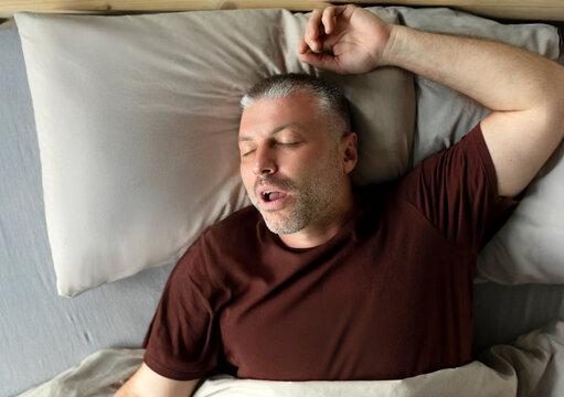 Middle aged man sleeping and snoring, lying with closed eyes in comfortable bed in bedroom at home, above view. Male sleep, recreation and wellness