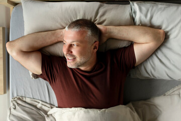 Fototapeta premium Happy middle aged man waking up and enjoying good morning lying in comfortable bed with hands behind head, above view