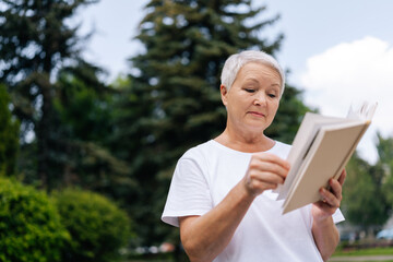 Low-angle view of gray-haired senior woman reading book in city park. Beautiful lady admiring green garden sitting on bench in summer day. Serene stylish pensioner holding literature resting outdoors