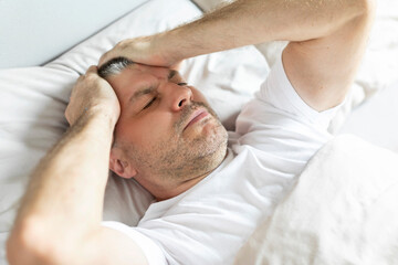 Middle aged man waking up in the morning with headache, suffering migraine, lying in bed and touching head