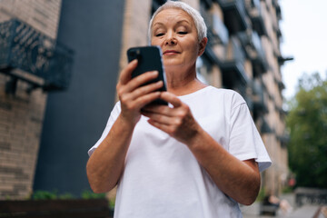 Low-angle view of gray-haired senior lady using smartphone texting message standing on urban street on summertime. Middle aged woman grandma hold phone typing sms enjoying communication in mobile app.