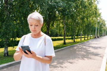 Pretty senior aged woman typing on smartphone while walking in city park. Beautiful gray-haired older woman using tapping mobile phone, smiling enjoying online communication.