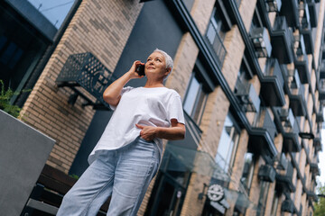 Low-angle view of beautiful mature grey-haired smiling woman in white clothes standing on fresh air talking on phone laughing speaking. Fashionable elderly female communicating using smartphone.