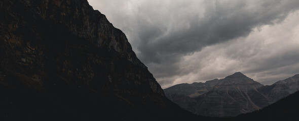 view of a rocky mountain face in the shadow with a mountain peak in the distance and heavy clouds in the sky