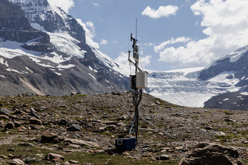 A weather monitoring station perched on rocky terrain in the foreground, with a vast glacier and towering mountain range seen beyond under a bright, clear sky.