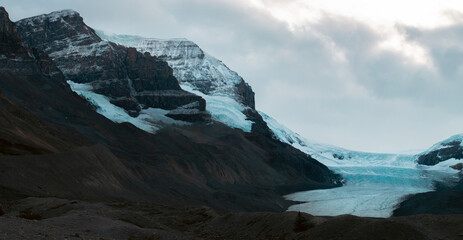 A vast glacier encased in snow stretches between two rugged mountains, embodying the harsh yet splendid beauty of the alpine environment and the sheer power of nature.