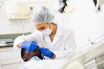 Naklejka premium Professional asian female dentist performing teeth treatment to adult african american man in dental clinic..