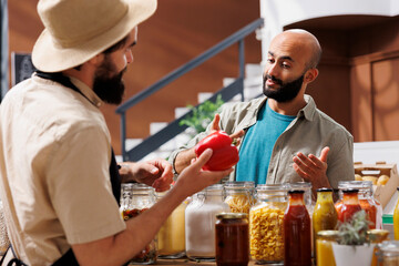 Middle eastern customer inquires about red bell pepper that a male storekeeper with a hat is holding. Vendor and client interacting over group of various glass jars and bottles in grocery store