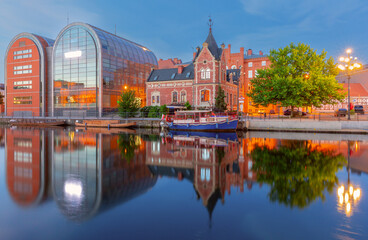 Fototapeta premium View of the Bydgoszcz waterfront featuring a modern glass building and a historic red brick structure, with reflections in the Brda River in Poland