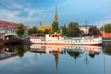 Obraz premium View of a church and boats reflected in the calm water of the Brda River at dawn in Bydgoszcz, Poland