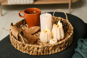 Wicker basket with burning candles in shape of pumpkins and tea cup on pouf at home, closeup